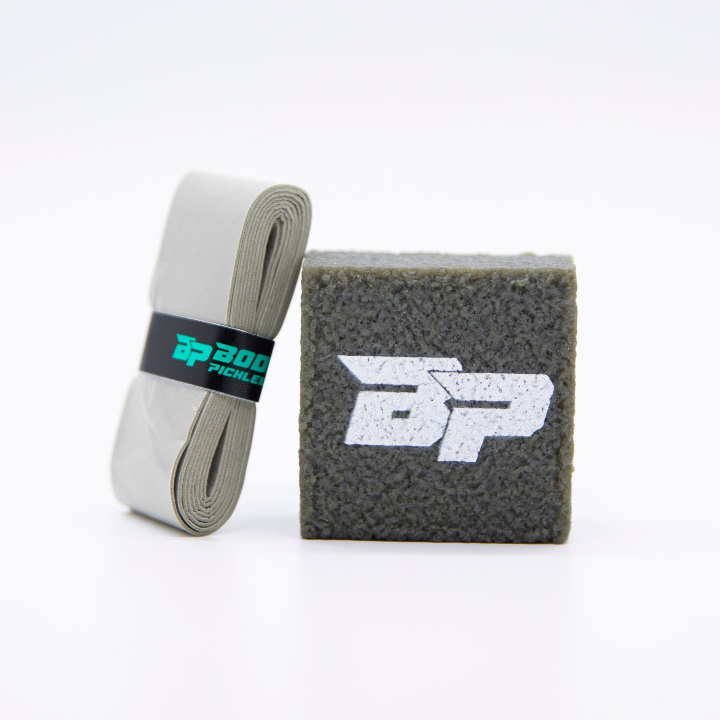 A rolled gray grip tape with a black label is beside a textured gray PROeraser™ pickleball paddle cleaner cube by Bodhi Performance, featuring a white BP logo on the front. Both are displayed against a plain white background.