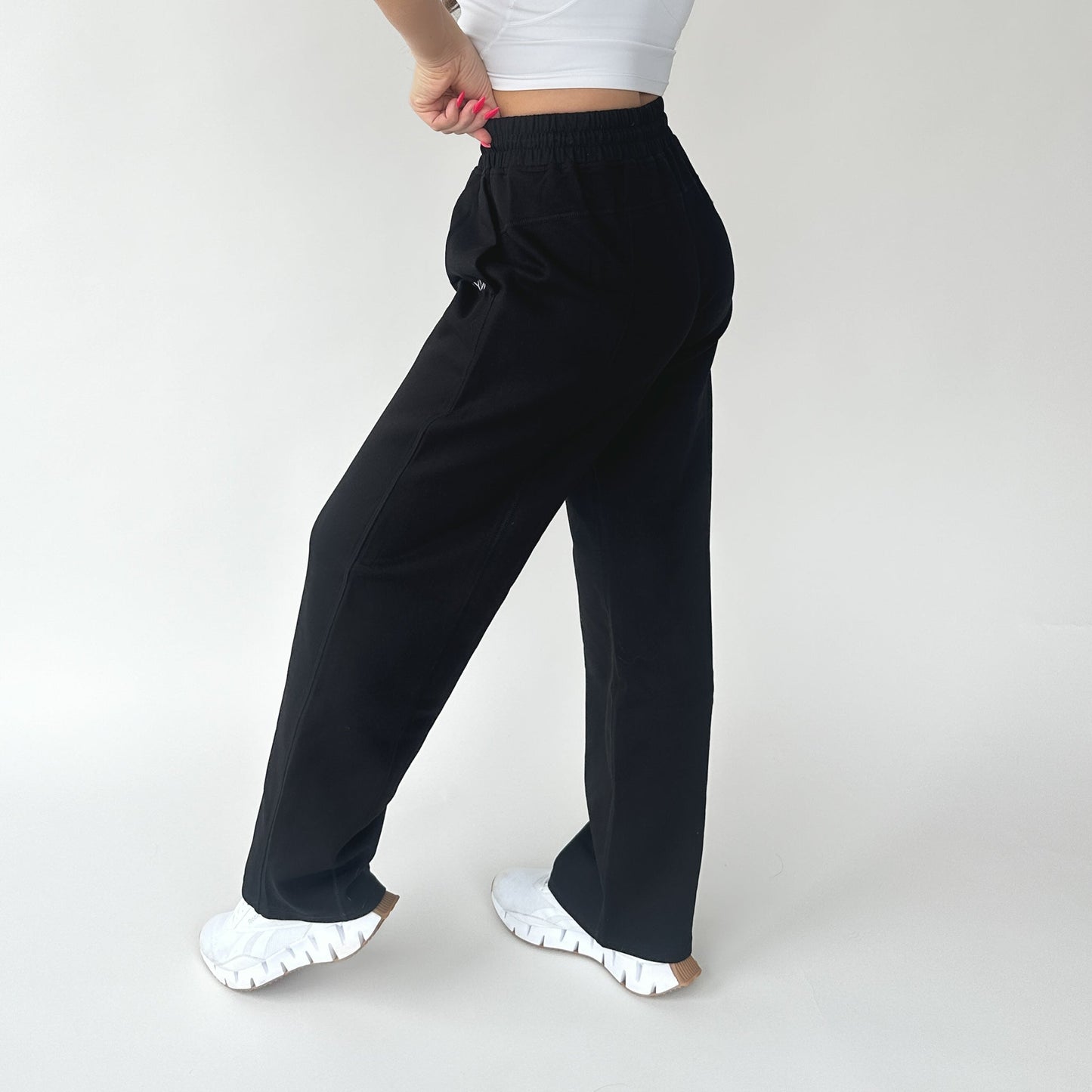 A person poses against a white background in DYM Athletics' Relax Wide Leg Sweatpant w/ Cinch Drawstring, paired with a fitted white crop top and sneakers. The side and back of the loungewear set are shown, with one arm bent and manicured nails visible.