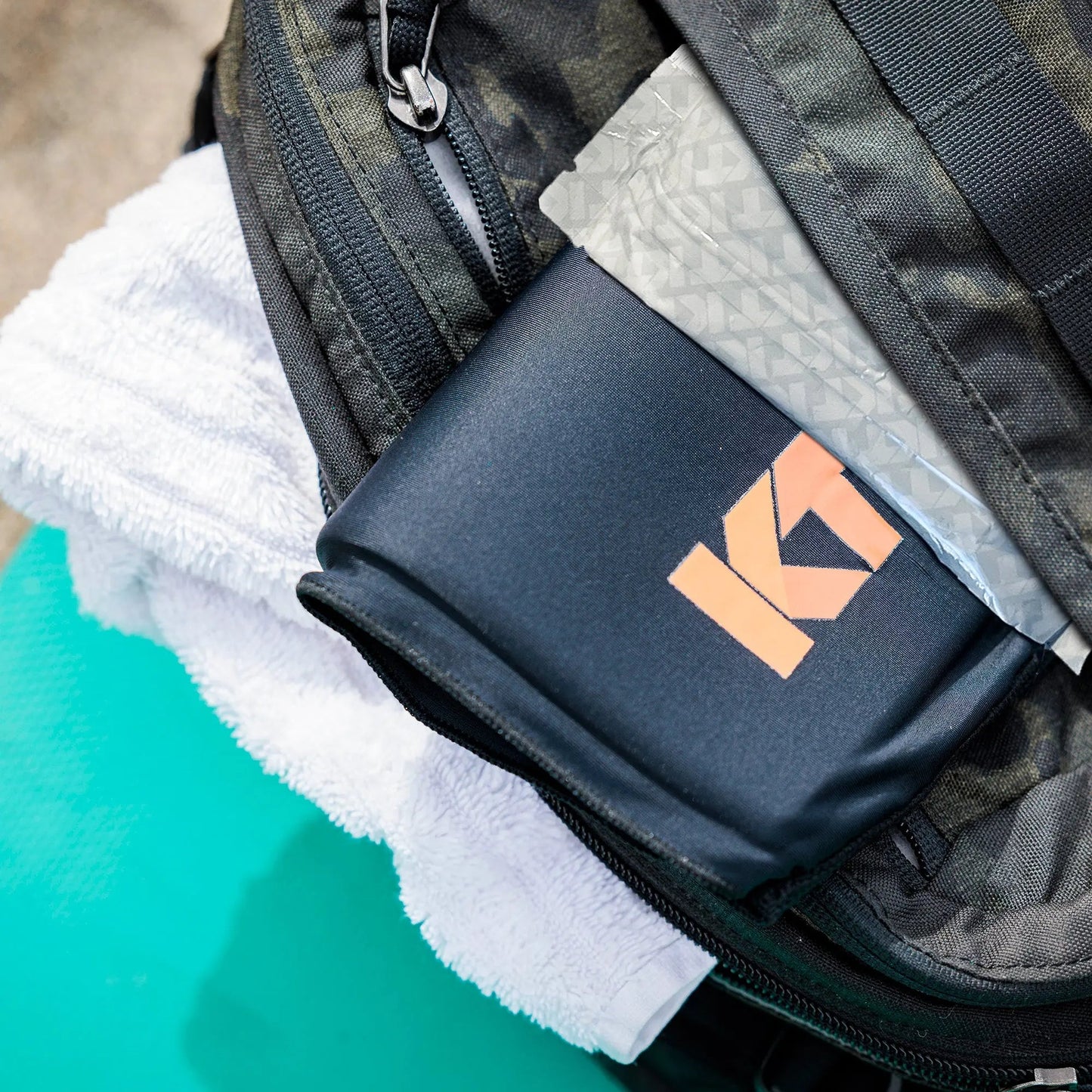A close-up of a black camo bag, partially unzipped to show a folded white towel, the KT Tape KT Health Ice Sleeve in light gray for cold therapy or sore joints, and a black item with the KT logo. The bag is on a turquoise surface.
