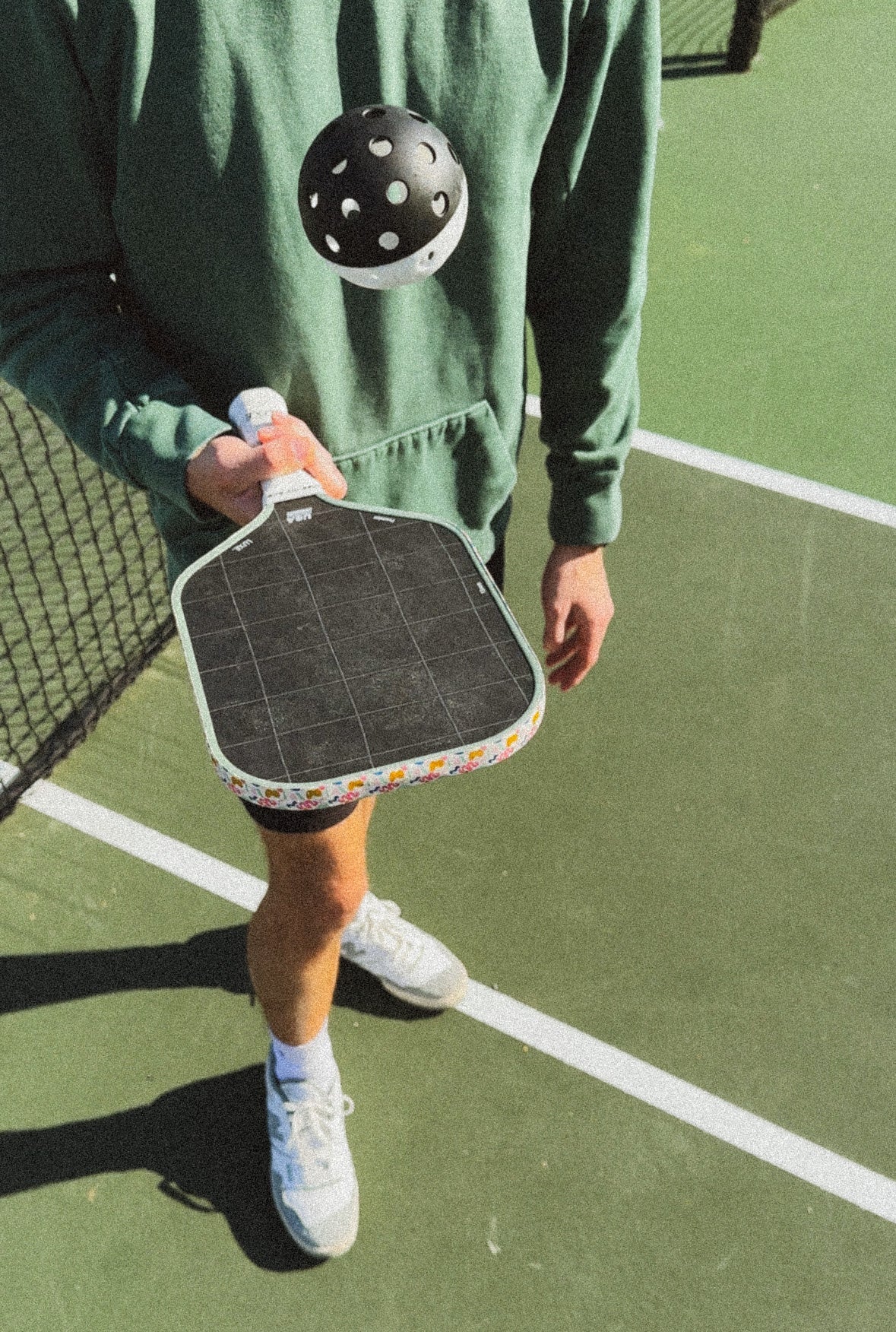 A person in green shorts stands on an outdoor pickleball court with green and white lines, holding the LUXE Pickleball Premier raw carbon fiber paddle and tossing a black perforated ball. Only their lower body and hand are visible.