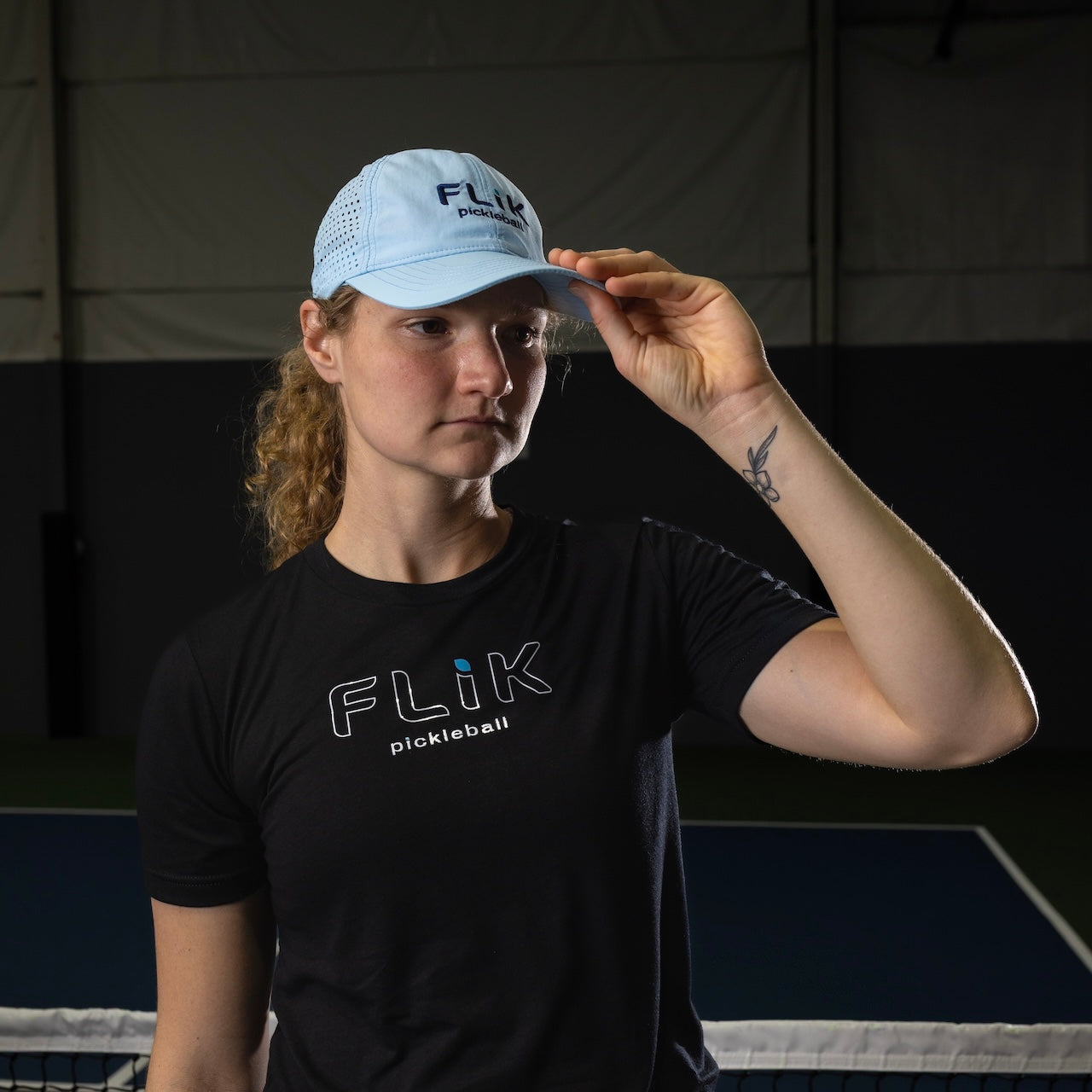 On a dimly lit indoor pickleball court, a woman in a light blue body helix FLiK Pickleball Hat and black FLiK shirt adjusts her cap, her brown curly hair tied back and a tattoo visible on her left forearm.