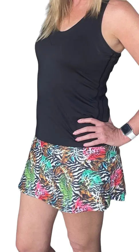 A woman poses in the Runway Athletics Nola Go Wild Court Skirt, a vibrant animal-print A-line tennis skirt with zebra and leopard patterns, styled with a sleeveless black top, smartwatch, and moisture-wicking under-short.