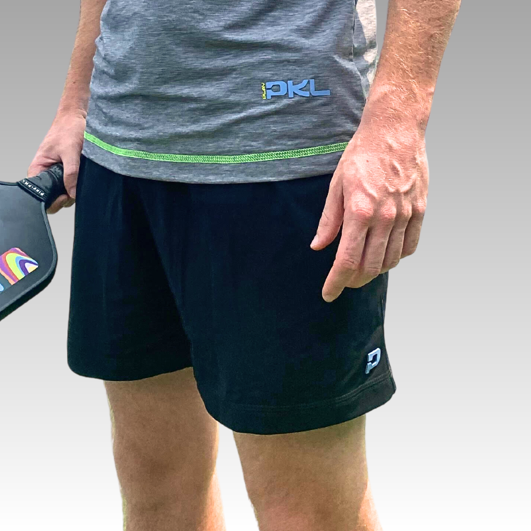 A person in a gray “PKL” shirt and black Playly Store Kelter Shorts is holding a black paddle with a colorful logo, standing against a plain gray background. Only the torso and legs are visible.