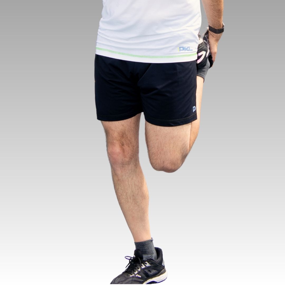 A person wearing a white shirt and Playly Store Kelter Shorts with black socks and sneakers stands on one leg, stretching the front thigh. The background fades smoothly from light gray to white; face is not visible.