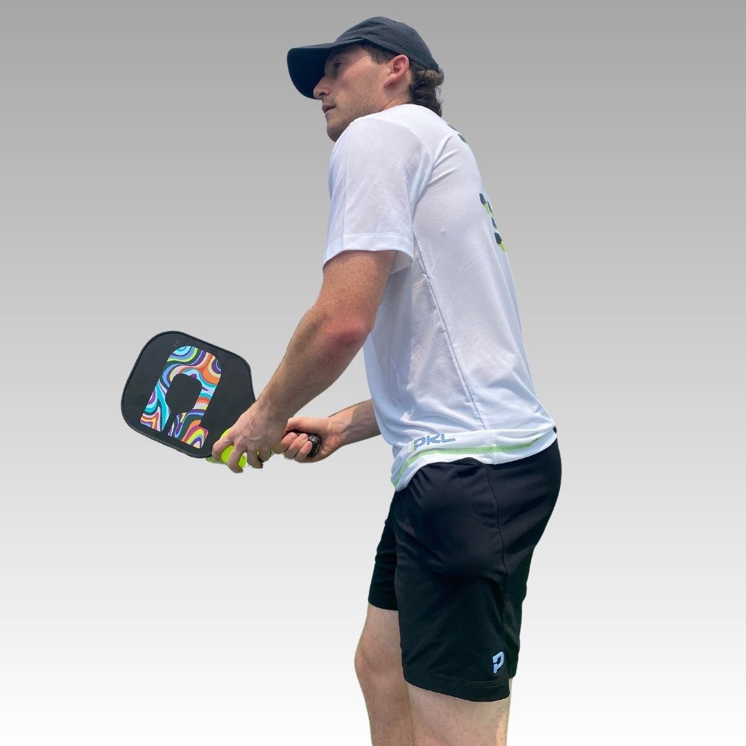 A man in a white shirt, black cap, and Playly Store Kelter Short made with SofKnit fabric holds a colorful pickleball paddle. Against a light gray background, he stands focused and ready for play.
