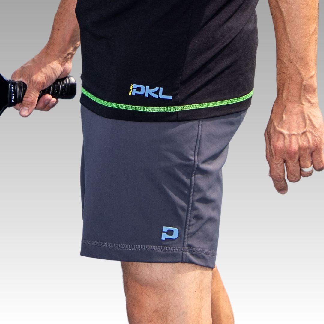 A close-up features a person wearing Playly Store’s Kelter Short in gray SofKnit fabric and a black tee with “PKL” in blue and yellow. They hold a pickleball paddle, with a wedding band on their left hand, against a plain gray background.
