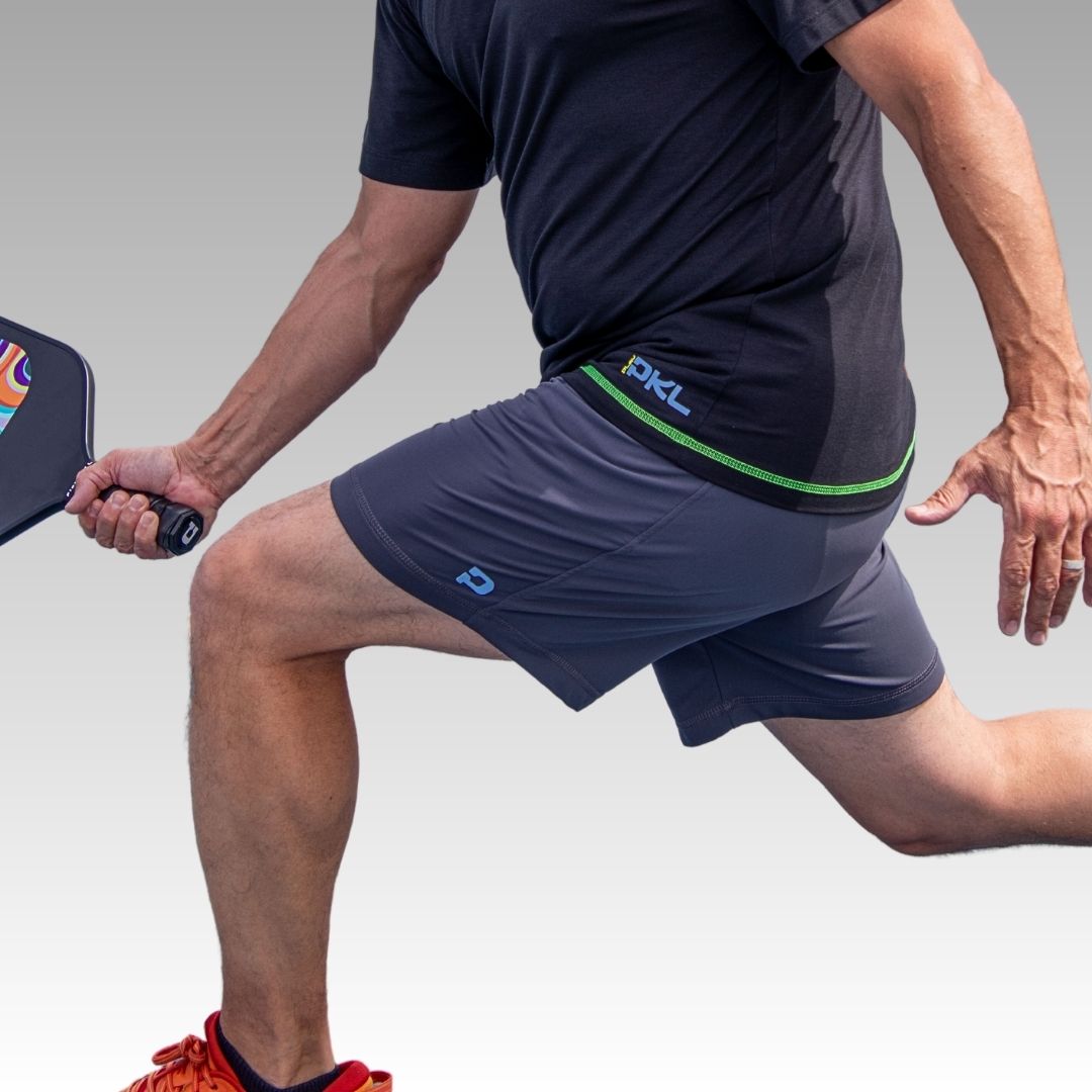 A person in a black shirt and gray Kelter Short from Playly Store lunges forward with a paddle in hand, their torso and legs shown against a smooth white-to-light gray gradient background.