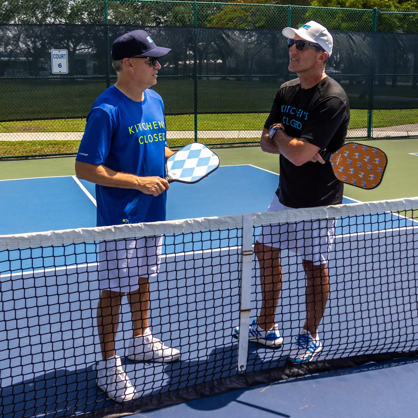 Two men wearing Swinton Pickleball Men's Kitchen's Closed Performance Shirt in Royal Blue, sunglasses, and hats stand on opposite sides of an outdoor pickleball net chatting before or after a sunny-day game.
