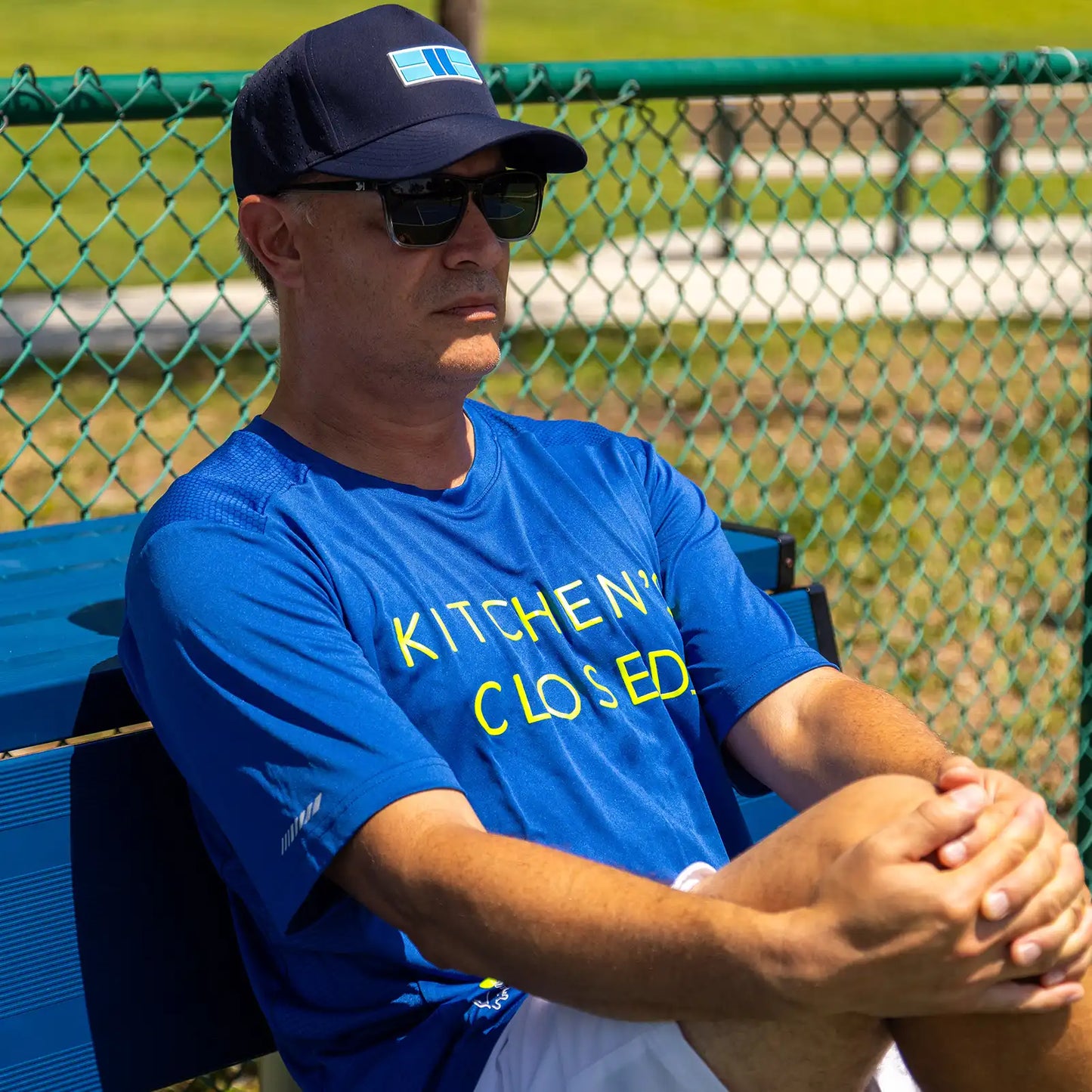 A man in sunglasses and a navy cap sits on a blue bench by a green fence, wearing the Swinton Pickleball Men's Kitchen's Closed Performance Shirt in Royal Blue. Yellow "KITCHEN CLOSED" text stands out; sunlight, grass, and pavement complete the scene.
