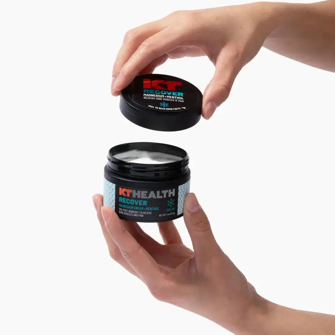 A person opens a black jar labeled KT Health Magnesium Creams by KT Tape with both hands, revealing the white magnesium cream inside. The lid is held above the open container against a plain white background.