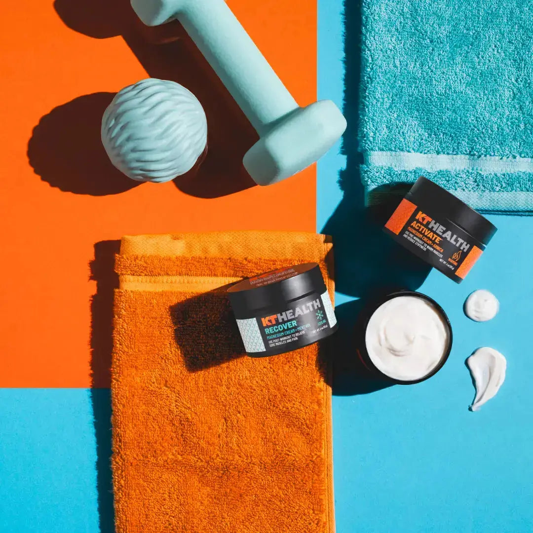 A blue dumbbell, textured massage ball, orange towel, and blue towel are arranged on an orange and blue background. Two KT Tape KT Health Magnesium Creams jars sit beside the towels; one jar is open to reveal white cream with swirls nearby.