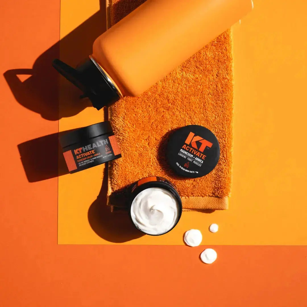 Flat lay of an orange water bottle, towel, KT Health Magnesium Creams by KT Tape (with some on the lid and next to it), and a small black KT Health Activate container, all on an orange background. Ideal for muscle recovery routines.