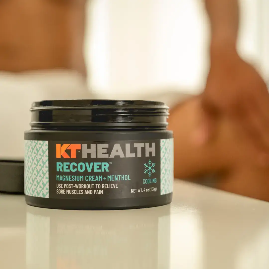 A close-up of a jar labeled “KT Health Magnesium Creams” from KT Tape sits on a white surface, highlighting this muscle relief cream for post-workout recovery. In the blurred background, a person touches their leg.