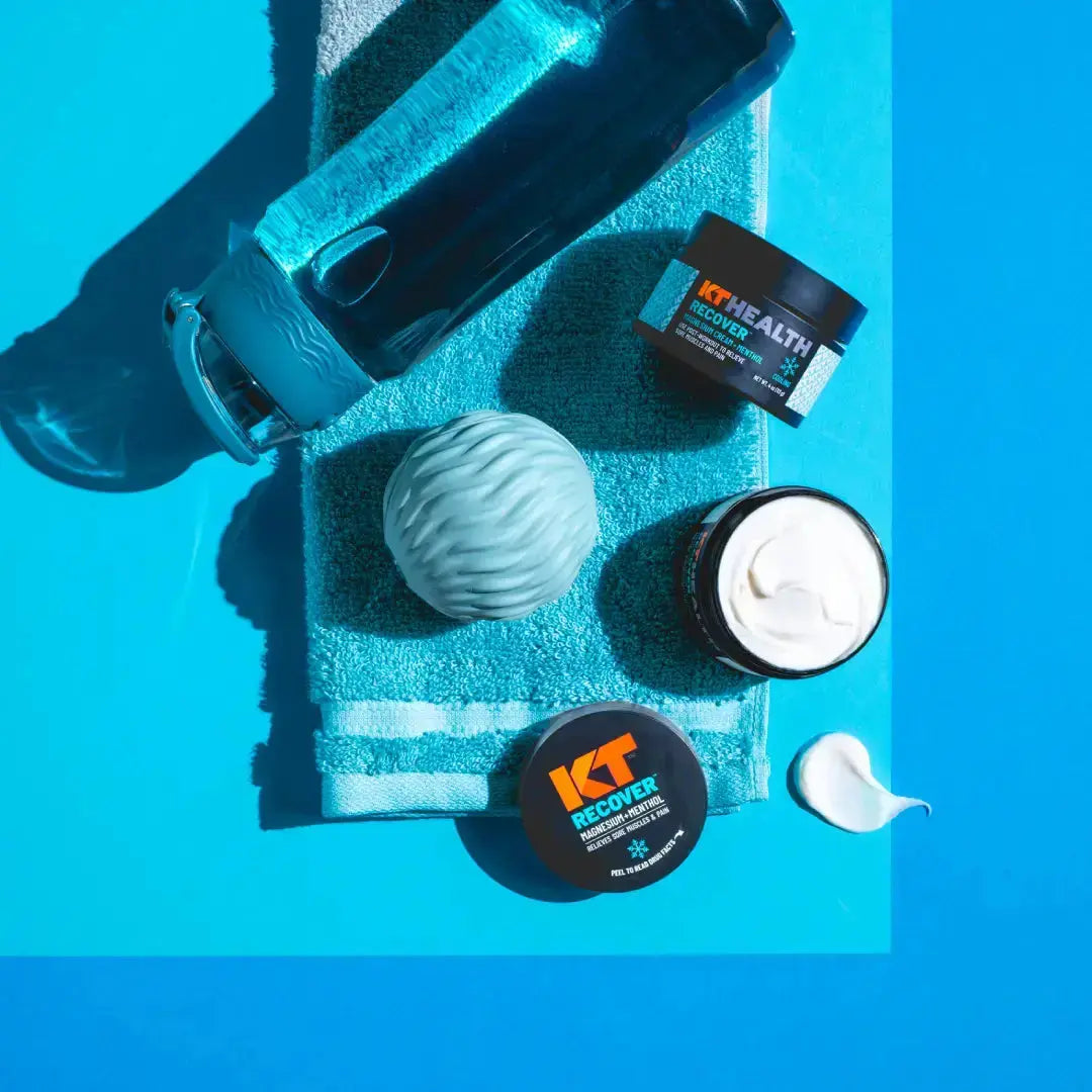 A blue towel, blue water bottle, textured blue massage ball, and two jars of KT Tape KT Health Magnesium Creams (one open with white cream) plus a dab of cream are displayed on a bright blue background under strong light.