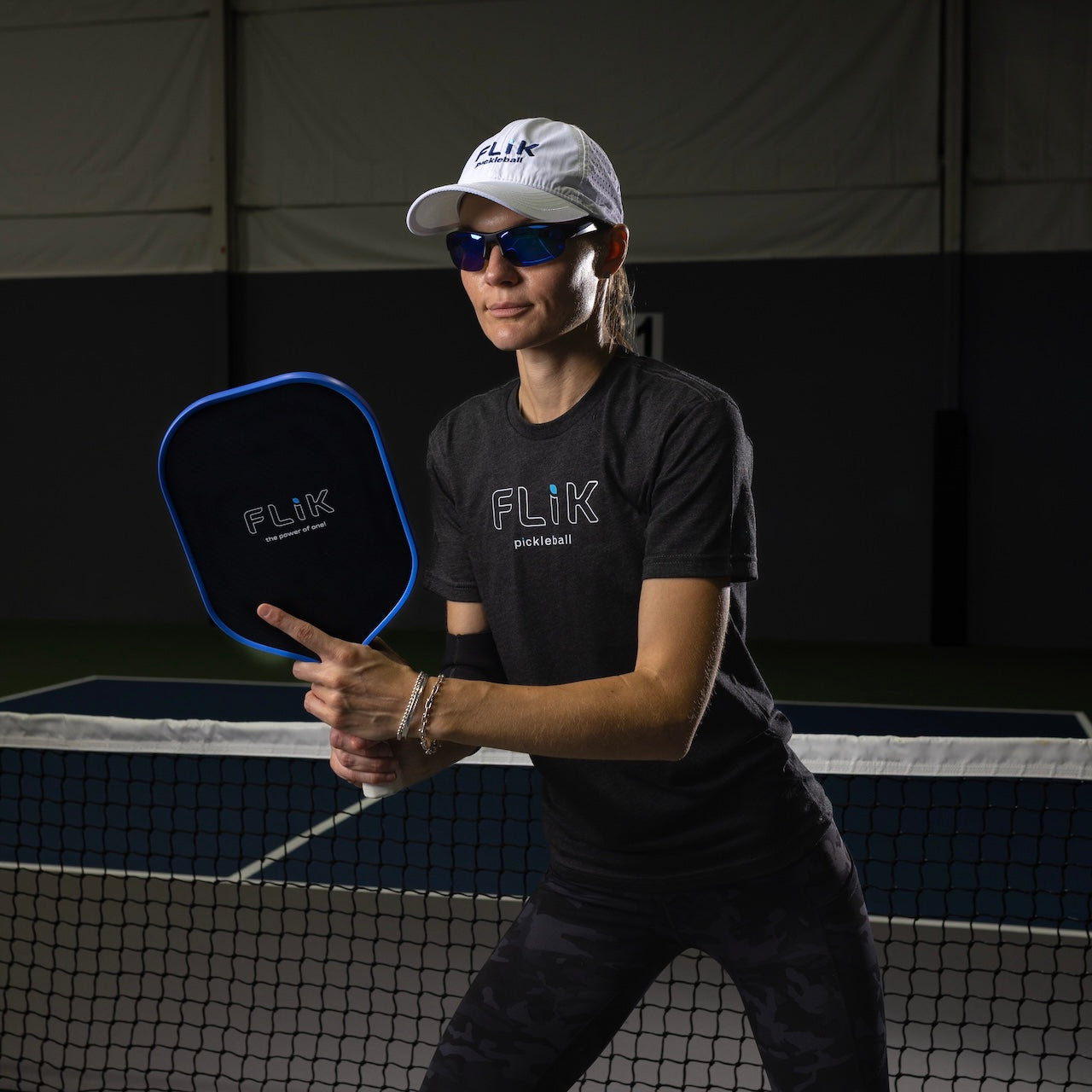 A woman in body helix Pickleball Glasses, a white hat, and a black FLiK Pickleball shirt stands ready with a paddle by the net on an indoor court. The background is dark and she appears focused in an athletic stance.