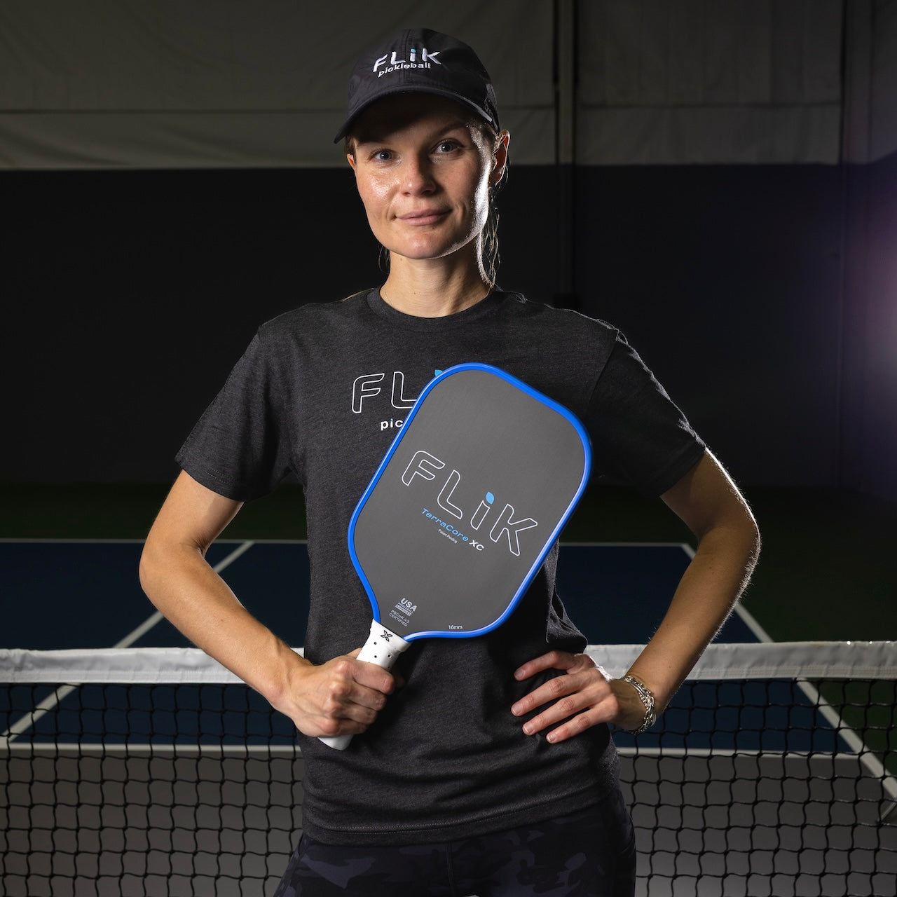 A woman in a black FLIK hat and shirt stands on a dimly lit indoor pickleball court, smiling with her left hand on her hip and holding a body helix FLiK F1 Elongated Pickleball Paddle—USAP Approved—in front of the net.