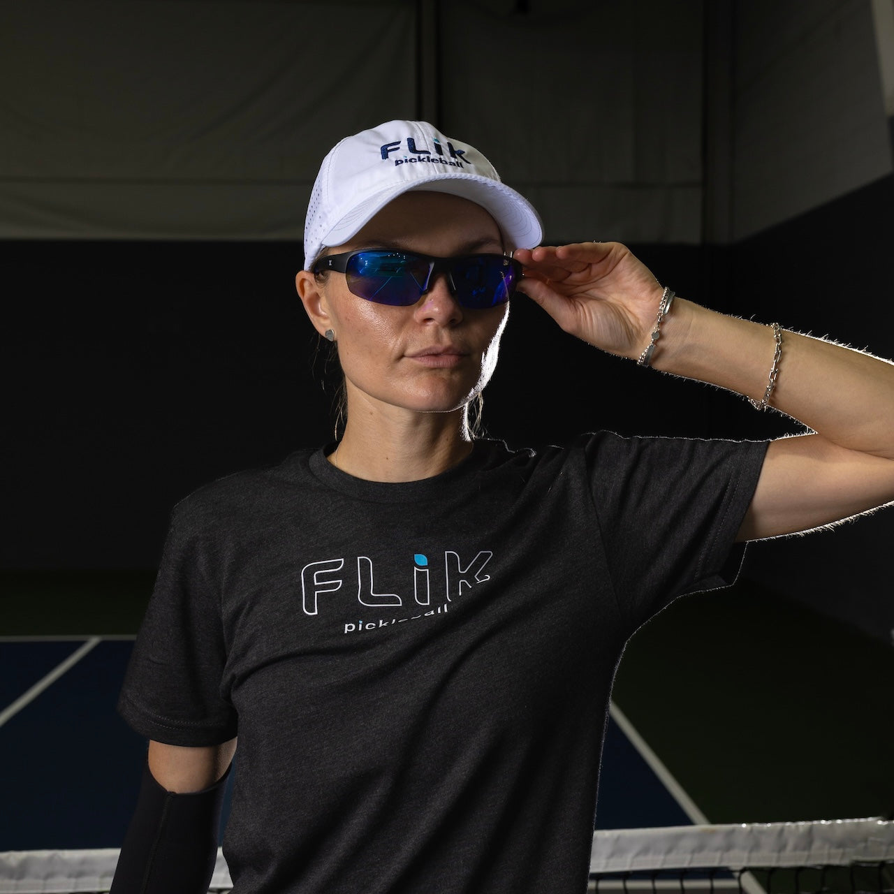 A woman stands on an indoor tennis court wearing a white body helix FLiK Pickleball Hat for sun protection, dark sunglasses, a FLiK pickleball t-shirt, and a bracelet. She holds her sunglasses, looking confidently at the camera in low light.
