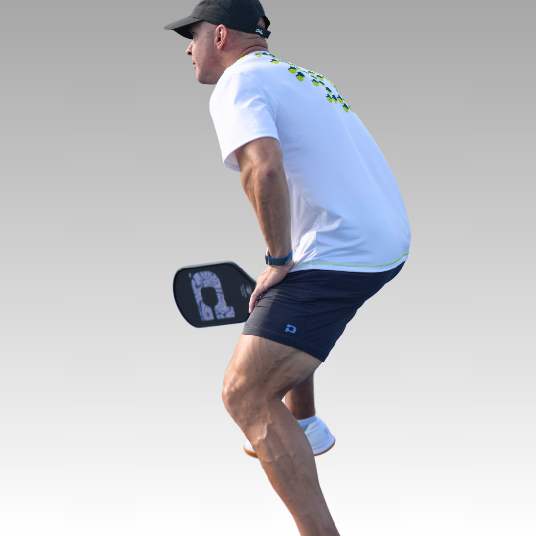 A man in a white shirt, black cap, and Kelter Shorts from Playly Store stands in an athletic stance, gripping his paddle with both hands against a light gray background.