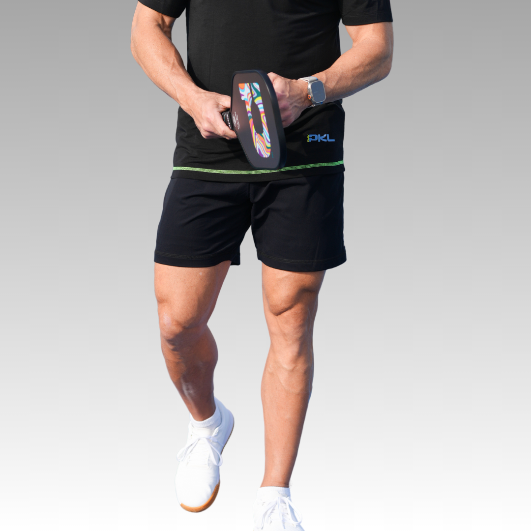 A person wearing a black shirt and Playly Store’s Kelter Short holds a colorful pickleball paddle at their waist. They stand ready to play, with white athletic shoes and a watch on their left wrist, against a plain gradient background.
