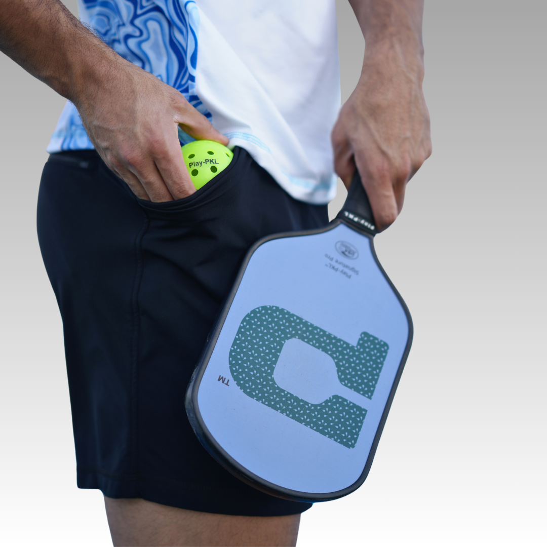 A person in Playly Store's Kelter Short and a white shirt with blue patterns holds a pickleball paddle, placing a yellow perforated ball into their pocket. The background is a plain light gradient.
