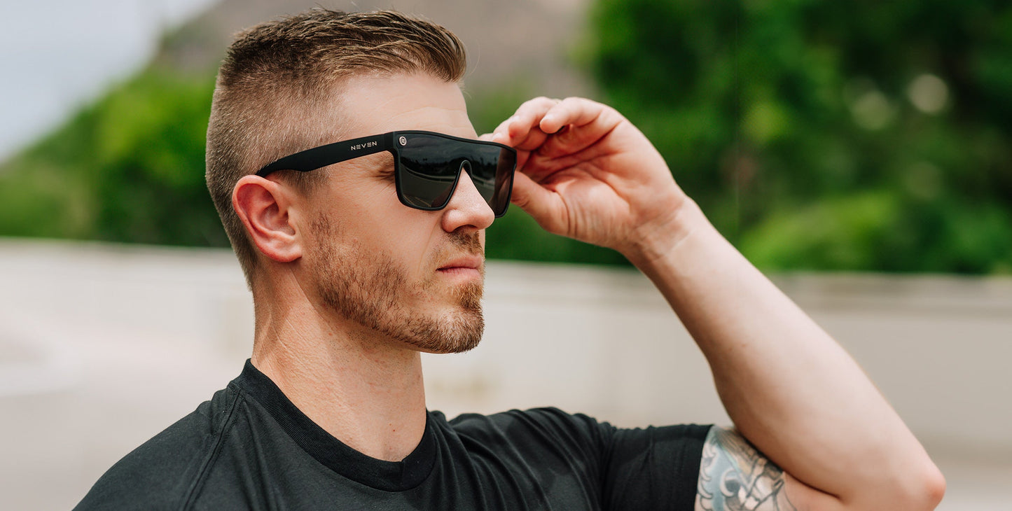 A man with short light brown hair and a trimmed beard wears Neven Eyewear Bold black polarized sunglasses and a black shirt. He is outdoors, adjusting his sunglasses with one hand; green trees blur in the background.