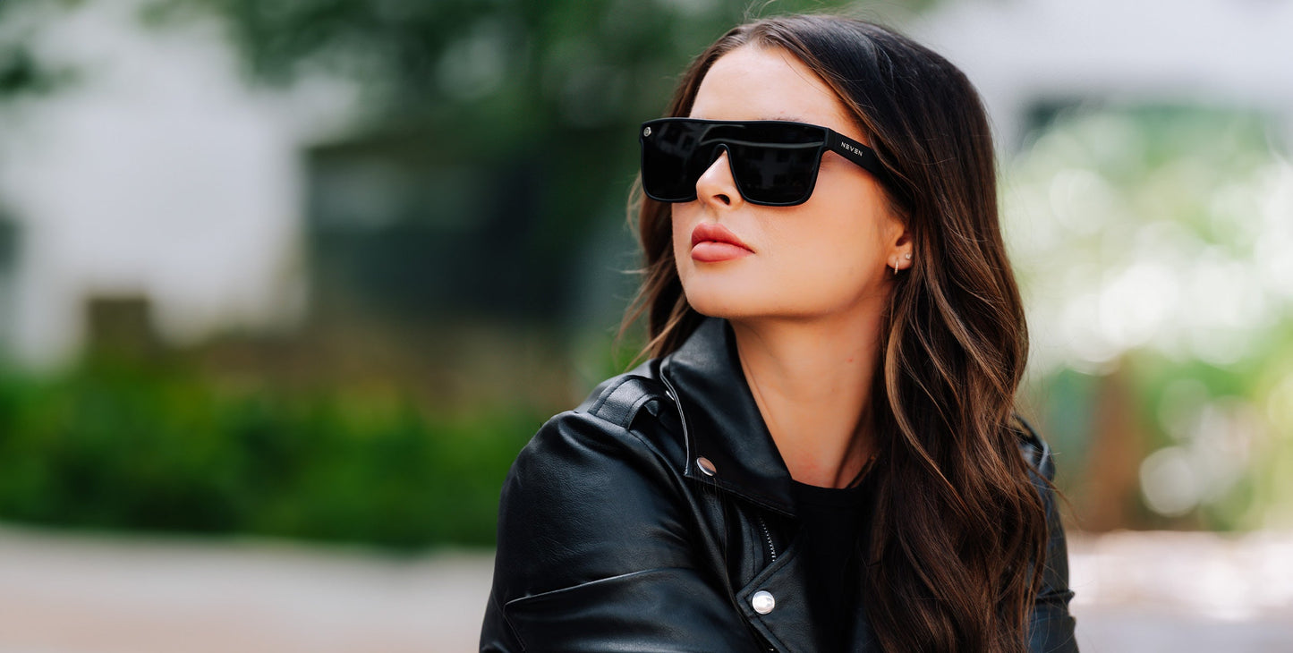 A woman with long brown wavy hair, wearing Neven Eyewear’s Bold black polarized sunglasses and a black leather jacket, looks to the side outdoors. The background is blurred with greenery and a white structure in natural, bright light.