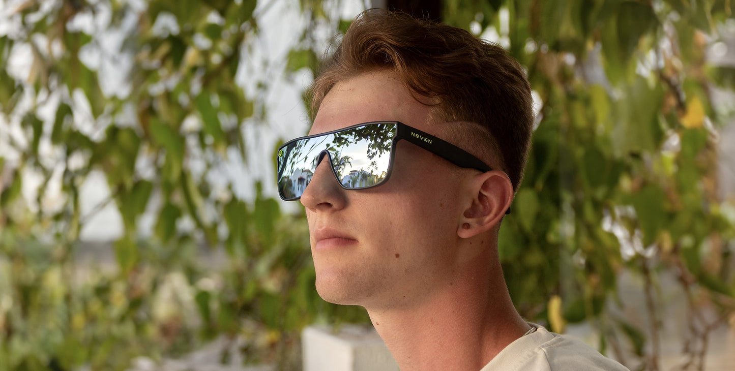 A young person with short, light brown hair wears Neven Eyewear’s CJ sunglasses featuring large, impact-resistant, polarized UV400 lenses. Calmly gazing upward in a white shirt, green foliage is softly reflected in the lenses.
