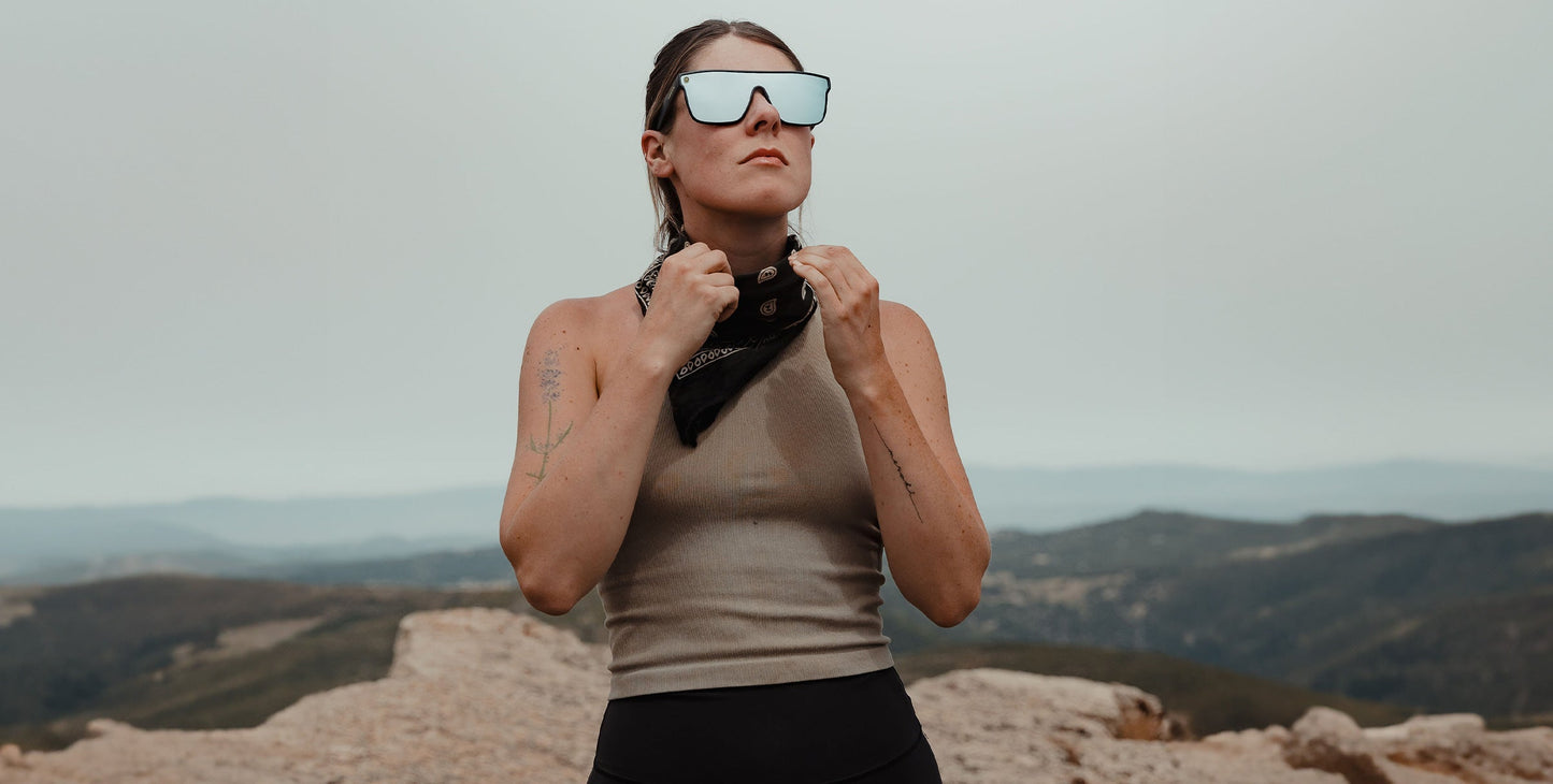 A woman wearing Neven Eyewear’s CJ large polarized sunglasses with UV400 protection and a sleeveless beige top stands on rocky terrain, tying a black bandana around her neck against hills and a hazy sky for an adventurous vibe.