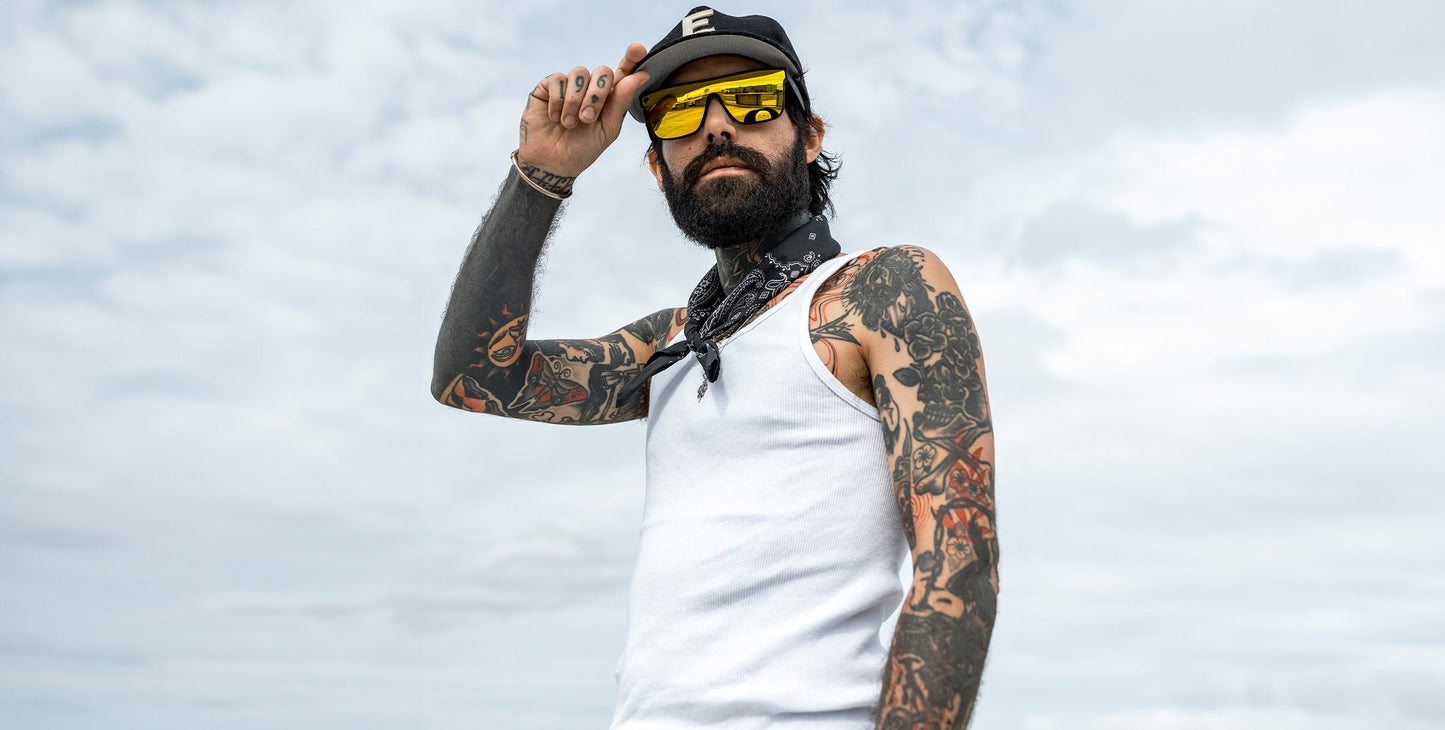 A tattooed man in a white tank, black cap, and black bandana stands outdoors under clouds, tilting his cap confidently. He wears Neven Eyewear CWM sunglasses.