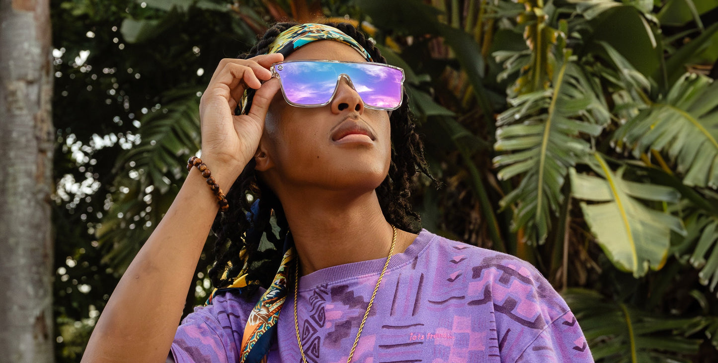 A person in a purple patterned shirt, colorful headscarf, and Neven Eyewear’s Fidelity polarized sunglasses stands outdoors among lush green tropical plants, gazing upward as sunlight highlights their face and accessories.