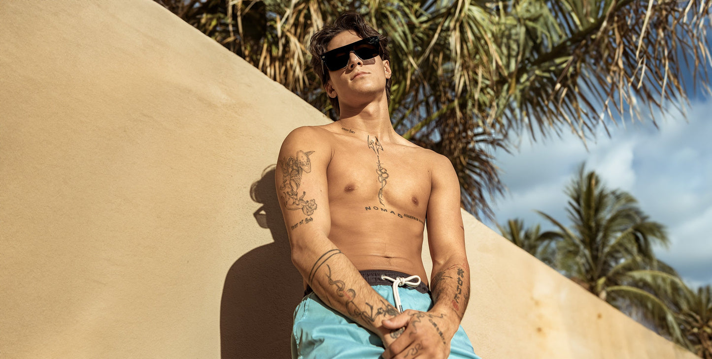 A young, shirtless man with tattoos wears Neven Eyewear’s Jordan sunglasses with UV400 protection, leaning against a tan wall under palm trees. He sports light blue swim trunks and gazes upward, relaxed, in the sunlight.