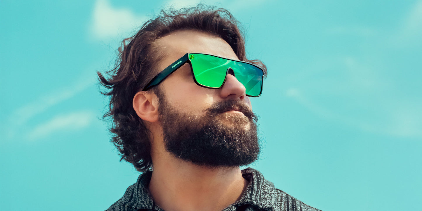 A bearded man with wavy brown hair sports Neven Eyewear's Lucky sunglasses—green polarized lenses and matte black frames—paired with a textured gray shirt, gazing confidently upward against a bright blue sky.