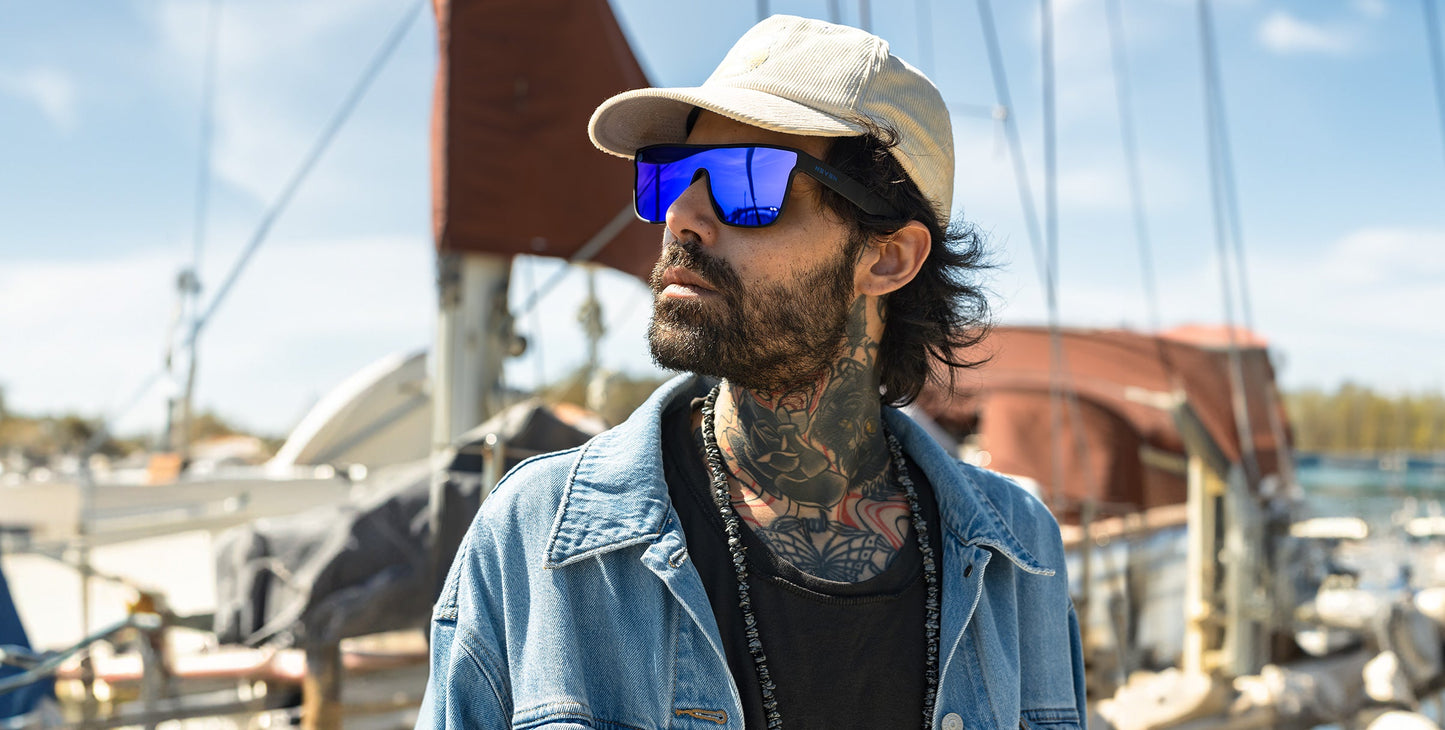 A man with tattoos on his neck and chest wears Neven Eyewear’s Midnight blue polarized sunglasses, a beige cap, and a denim jacket. He stands outdoors near sailboats under a sunny sky, looking to the side.