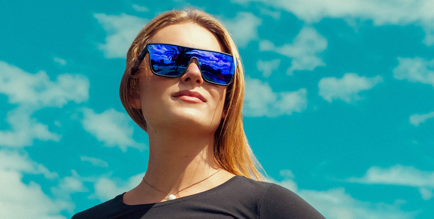 A woman with straight, light brown hair wears Neven Eyewear's Midnight sunglasses from the Legend collection and a black top, standing outdoors under a blue sky with white clouds, gazing confidently into the distance.