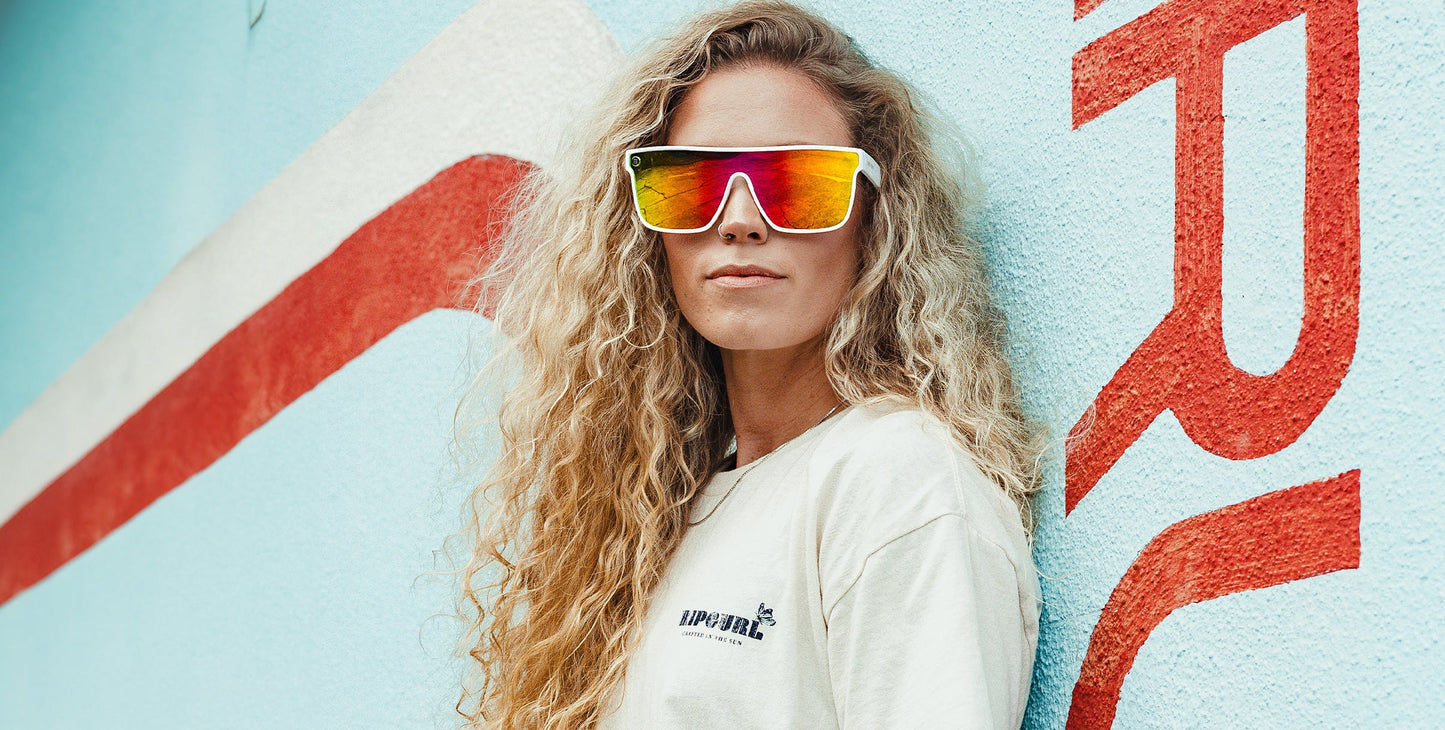A woman with long, curly blonde hair confidently models Neven Eyewear’s Muse sunglasses, featuring large reflective white frames with UV400 protection, paired with a cream t-shirt against a blue wall with abstract red and white designs.