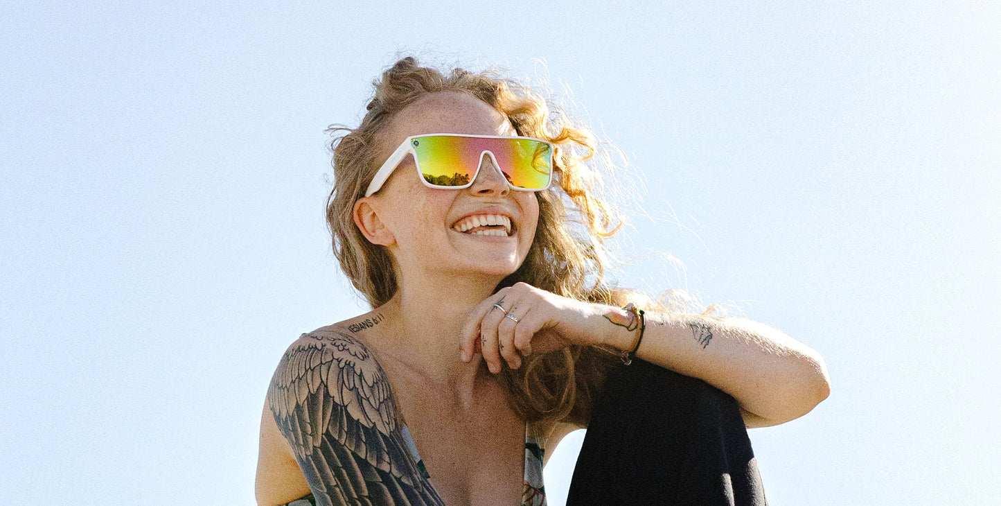 A smiling person with curly hair wears Neven Eyewear’s Muse polarized sunglasses, showcasing a wing tattoo on their shoulder while sitting casually outdoors against a blue sky, one arm on their knee and the other touching their chin.