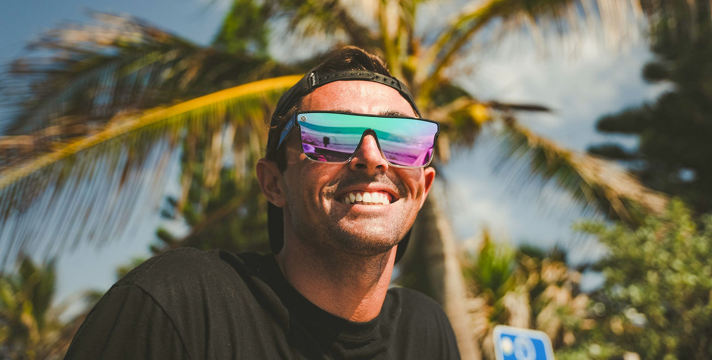 A man in a black t-shirt and backward cap smiles outdoors, wearing Neven Eyewear's Nomads polarized sunglasses (UV400 protection), which reflect a vibrant beach scene amid palm trees and blue sky, evoking a tropical vibe.