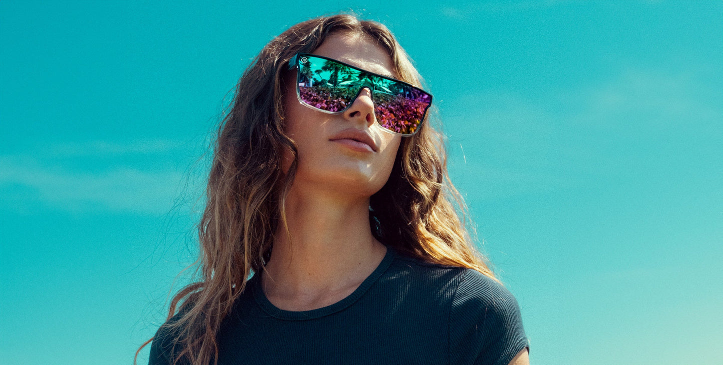 A woman with wavy hair wears a dark tee and large, colorful mirrored polarized Nomads sunglasses by Neven Eyewear. Reflections of people and trees gleam as she stands confidently under a blue sky, enjoying UV400 protection.