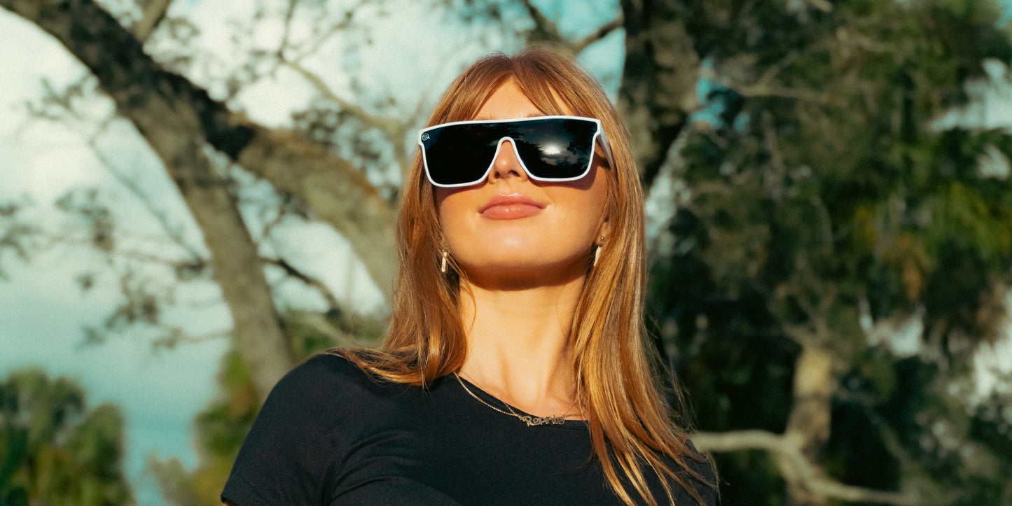 A woman with straight, light brown hair wears Permafrost sunglasses by Neven Eyewear and a black shirt. Outdoors, sunlight highlights her face as she smiles softly, with trees and a blue sky blurred in the background.