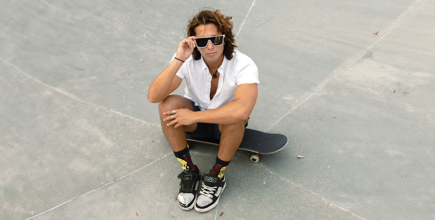 A young man with long hair sits on a skateboard, wearing a white shirt, black shorts, and Neven Eyewear's Permafrost sunglasses with polarized UV400 protection. He holds the minimalist frames, finishing his look with stylish flair.