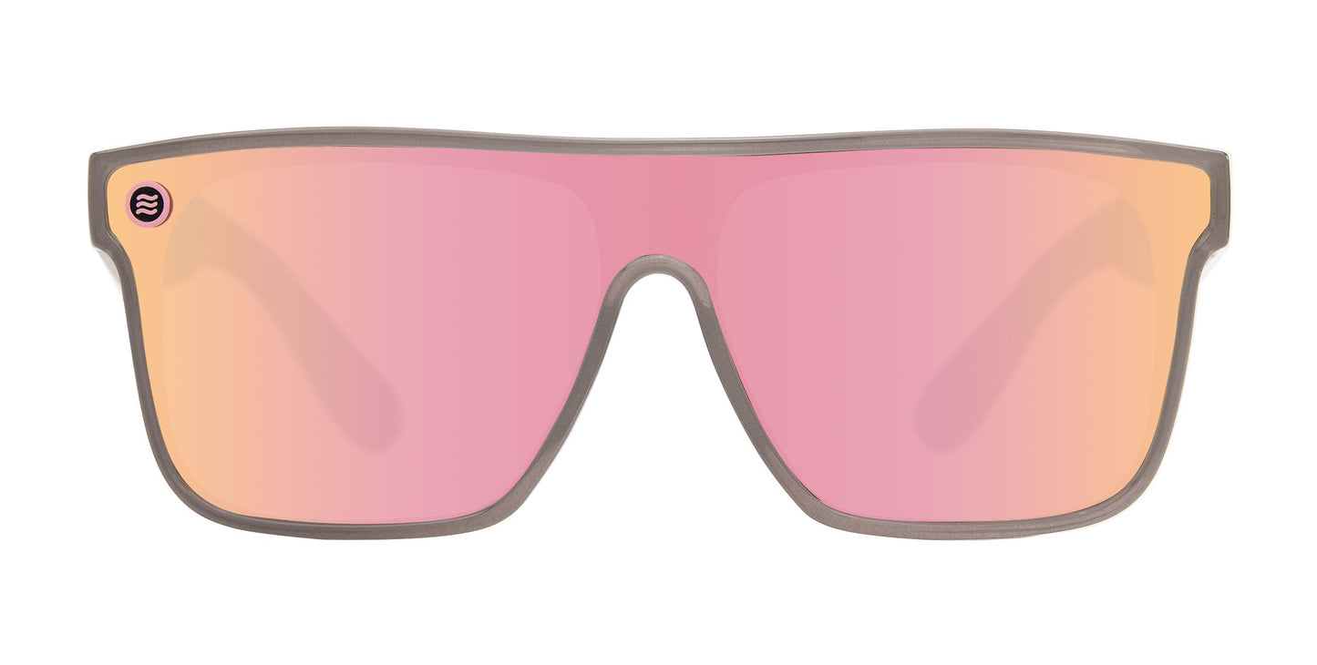 The Neven Eyewear Smooth Crush sunglasses feature a thin grayish frame, a pink-orange polarized shield lens, and a small purple circular logo on the upper left. Impact-resistant and stylish, they're set against a white background.