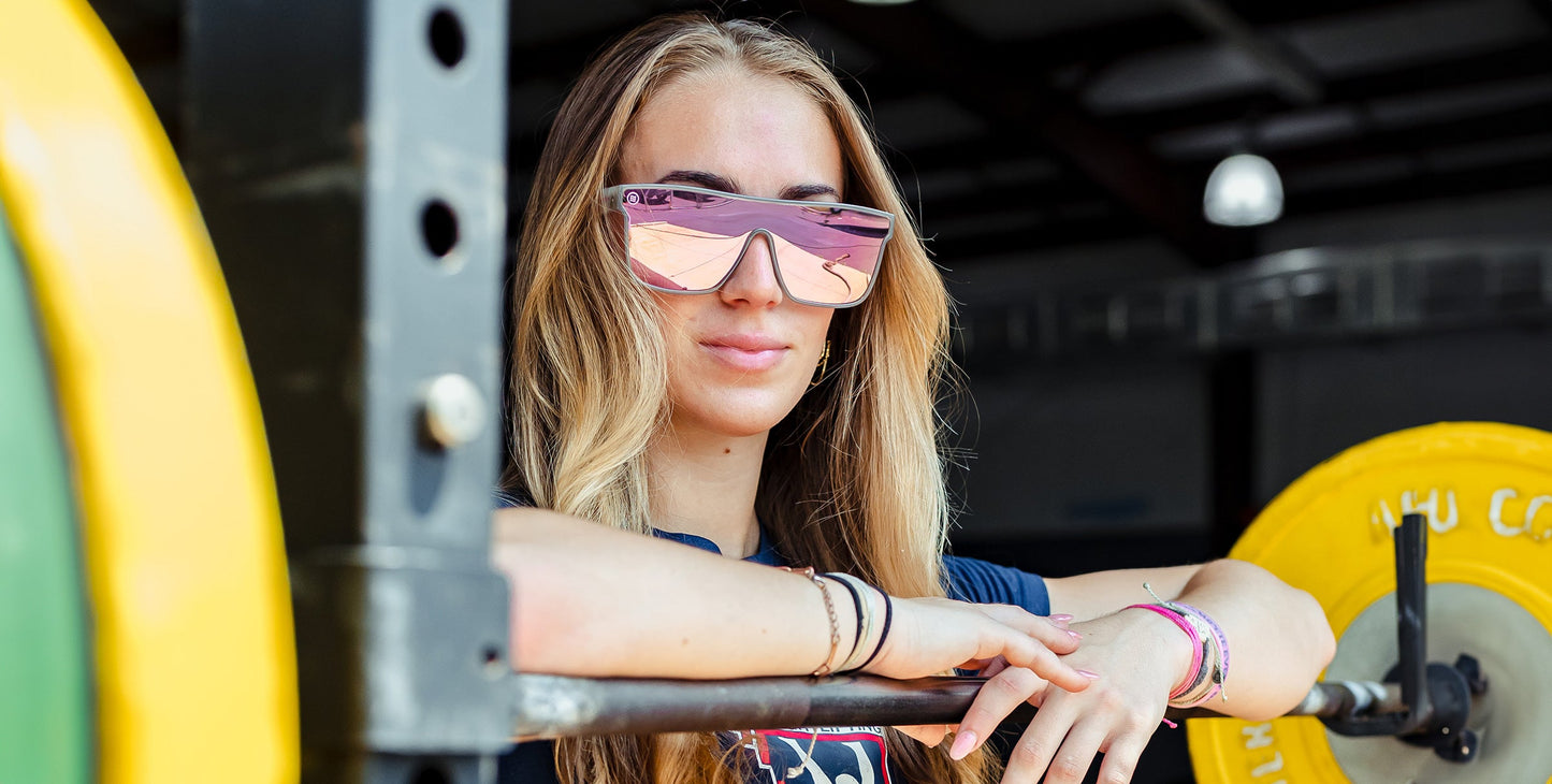 A woman with long blonde hair wears Neven Eyewear's Smooth Crush pink polarized sunglasses, a navy shirt, and bracelets as she leans on a barbell rack. Colorful weights stand out in the high-ceilinged, industrial-lit gym.
