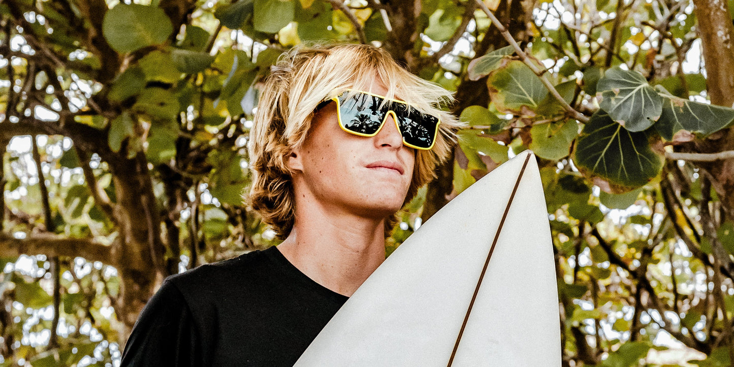 A young person with shaggy blond hair wears Neven Eyewear's The Swarm yellow sunglasses (UV400 protection) and a black shirt, holding a white surfboard outdoors in dappled sunlight with leafy trees in the background.