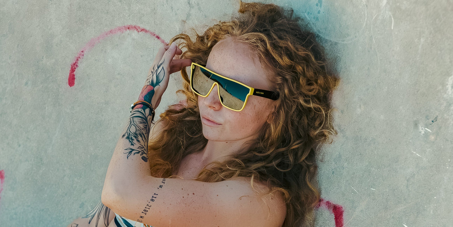 A person with long curly hair and tattooed arms lounges against a pink graffiti wall, wearing The Swarm sunglasses by Neven Eyewear with UV400 protection and a striped top, looking relaxed in the bright sunlight.