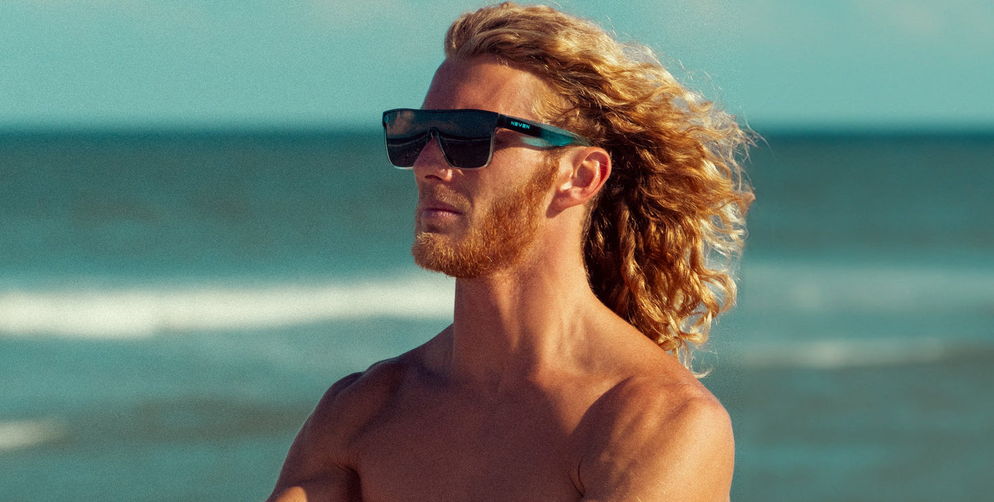 A shirtless, long-haired blond person with a reddish beard stands by the ocean in Neven Eyewear's Turnstile sunglasses featuring UV400 protection. They gaze into the distance as waves and blue sky suggest a sunny beach day.