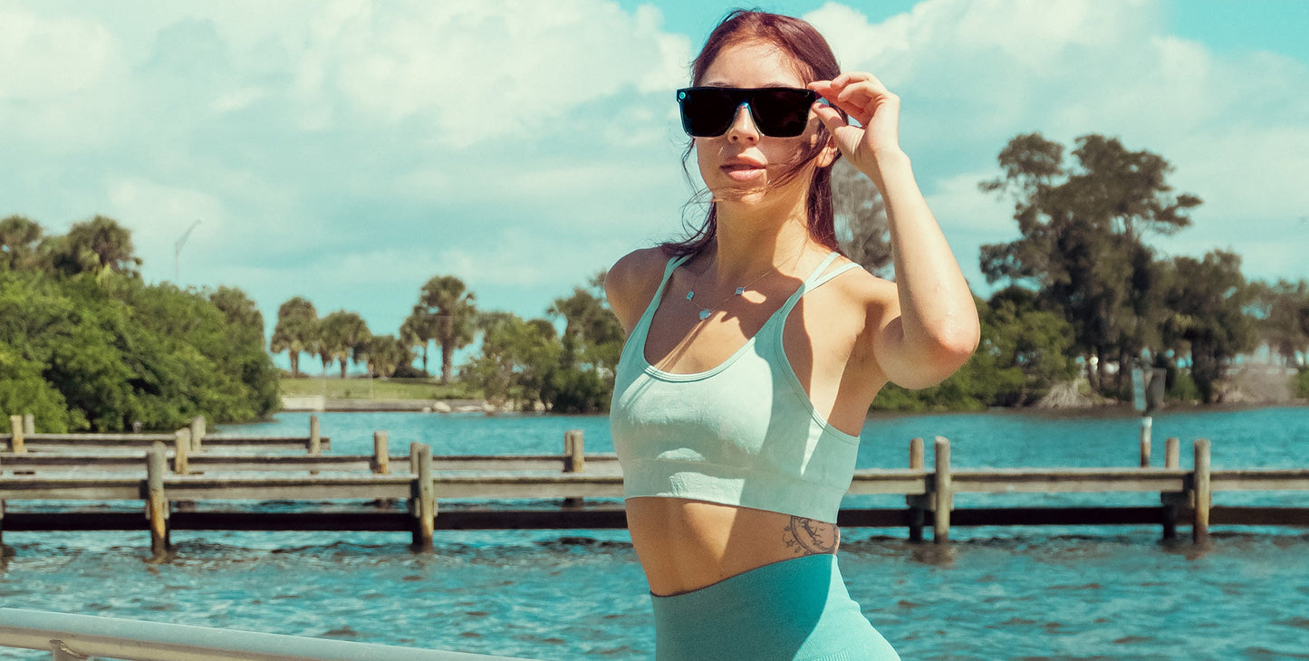 A woman with long reddish hair wears Neven Eyewear's Turnstile polarized sunglasses, a light blue sports bra, and leggings while standing outdoors by the water near trees and a wooden dock, looking at the camera.
