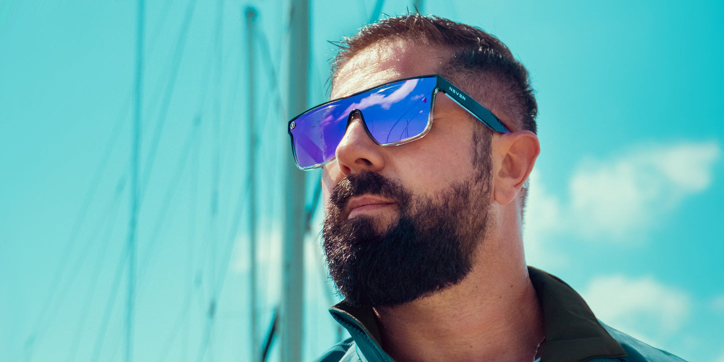 A bearded man with short dark hair wears Neven Eyewear's Nomads XL reflective blue polarized sunglasses and a teal jacket, standing outdoors on a sunny day. Blurred sailboat masts and a blue sky with scattered clouds are in the background.