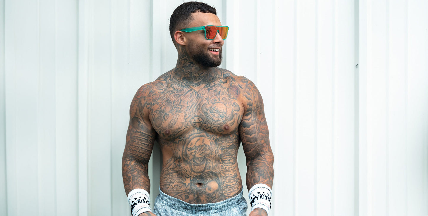 A muscular, tattooed man with a beard stands shirtless against a white corrugated wall, smiling to the side. He wears Neven Eyewear X hot pink polarized sunglasses with UV400 protection, white wristbands, and light blue shorts.