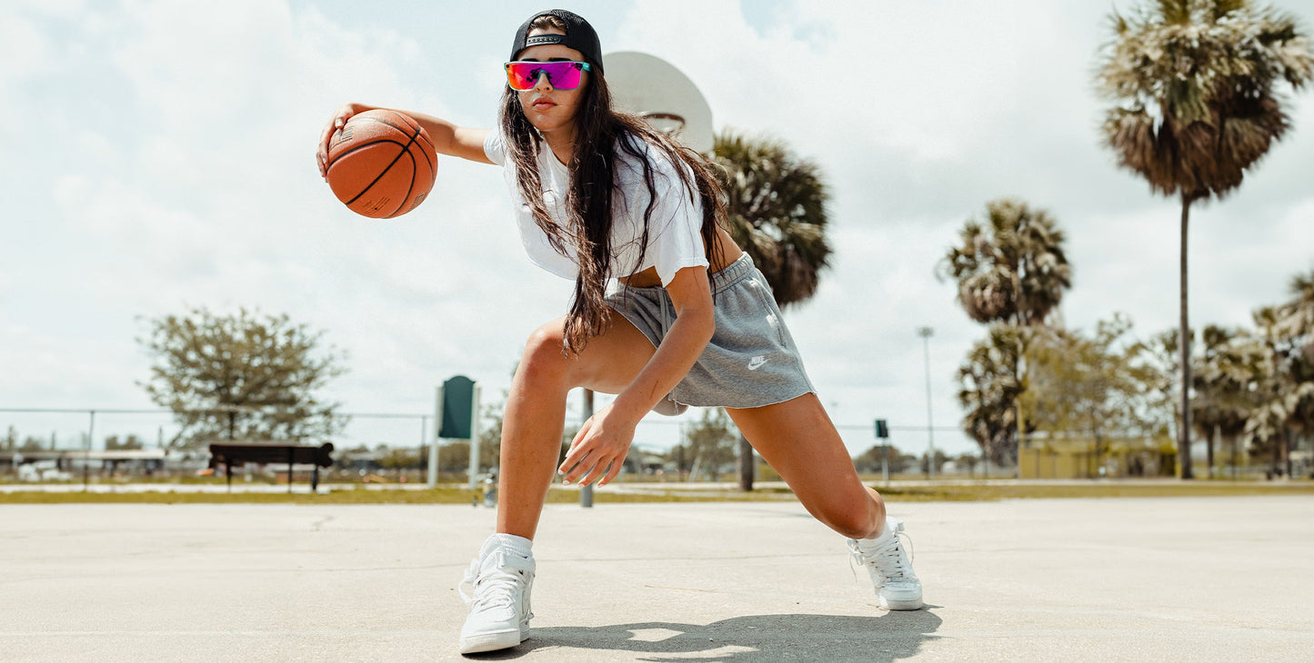 A young woman dribbles a basketball on an outdoor court, wearing Neven Eyewear’s X hot pink polarized sunglasses with UV400 protection, plus a black backward cap, crop top, and shorts under palm trees and the sunny sky.