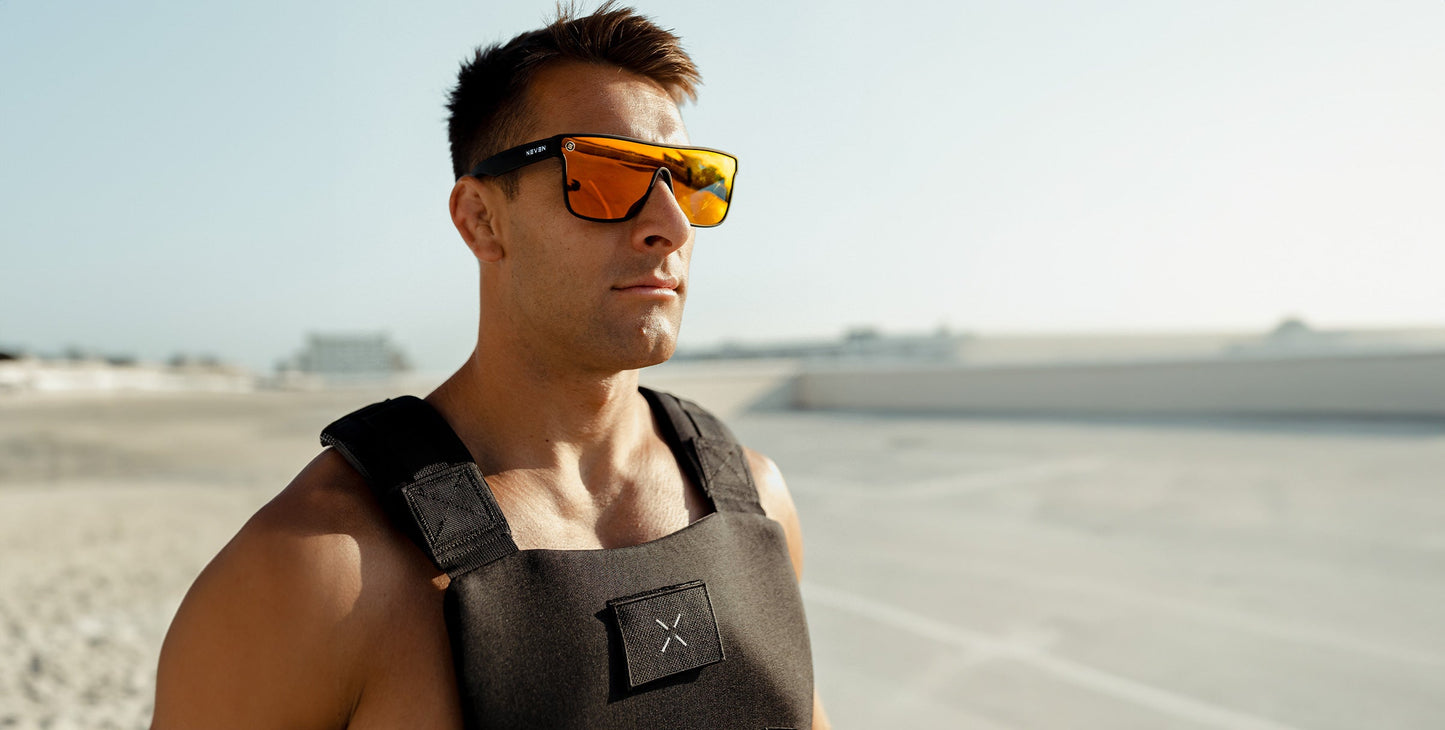 A muscular man stands confidently outdoors on a sunny day, wearing Neven Eyewear’s Zero Cool sunglasses with polarized UV400 protection and a weighted vest, set against a clear sky and blurred, sandy landscape.